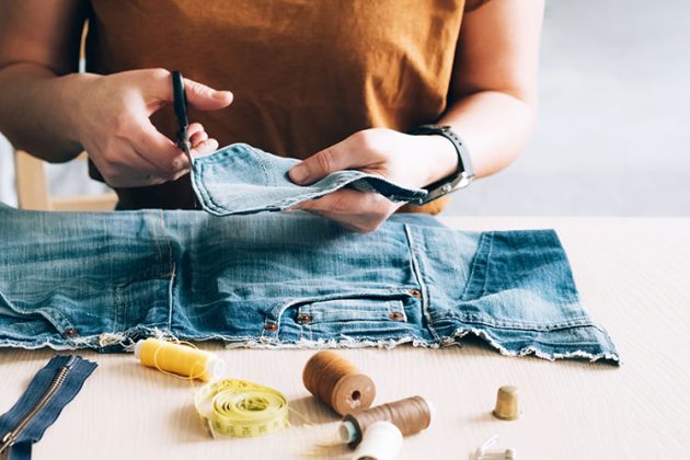 A woman repairs a pair of jeans on a table surrounded my fabric materials. The Case for Circular Fashion and Apparel in Vancouver