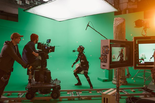 A film crew works on set in front of a green screen with lots of cameras and sound equipment. A man wears a motion capture suit